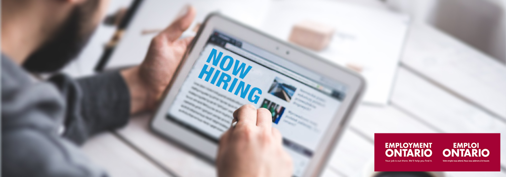 A man looks at a tablet with the words "Now Hiring" on the screen.